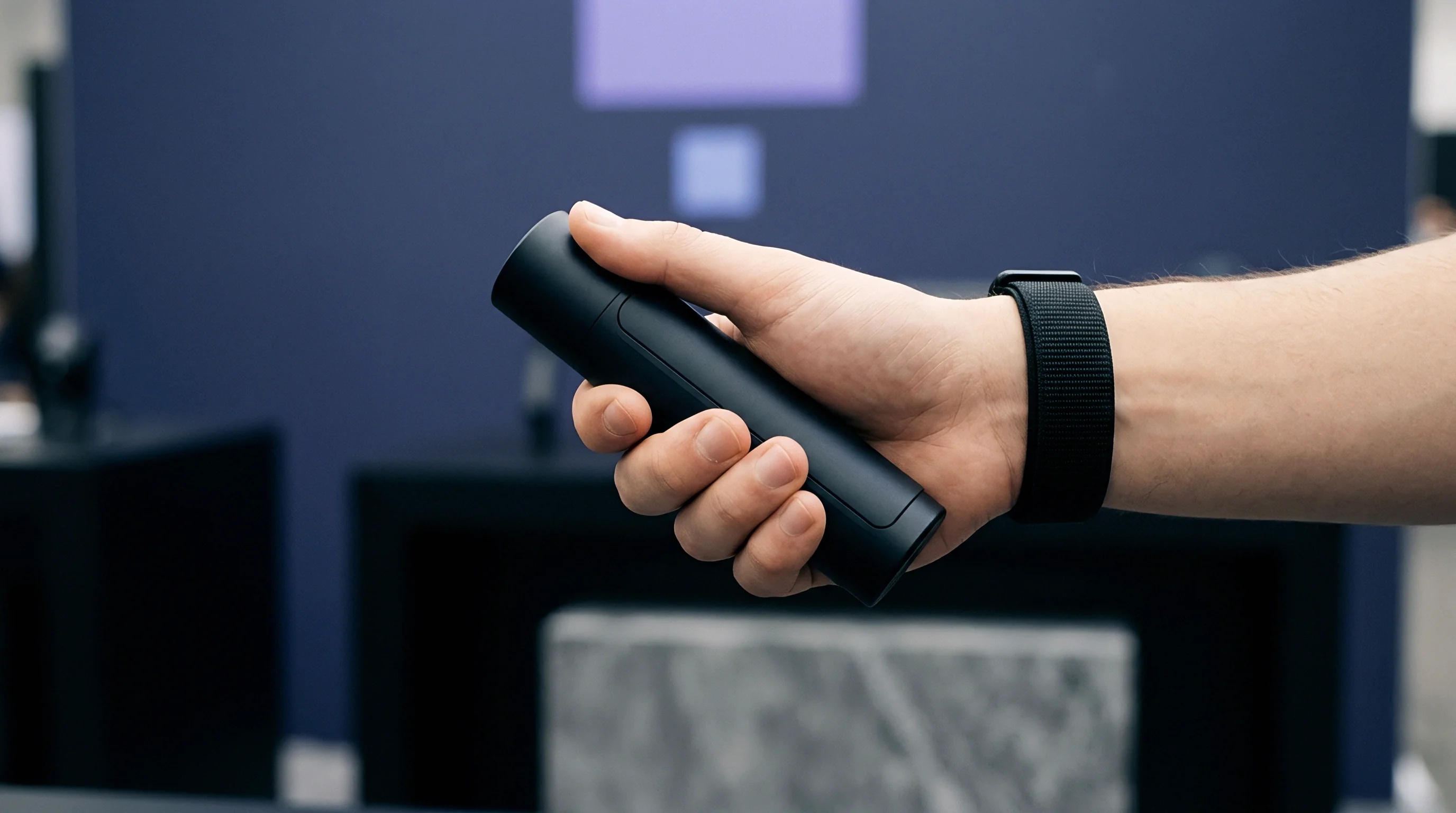 Fingers firmly grasp a matte dark cylindrical hardware prototype displayed inside a commercial trade exhibition space.
