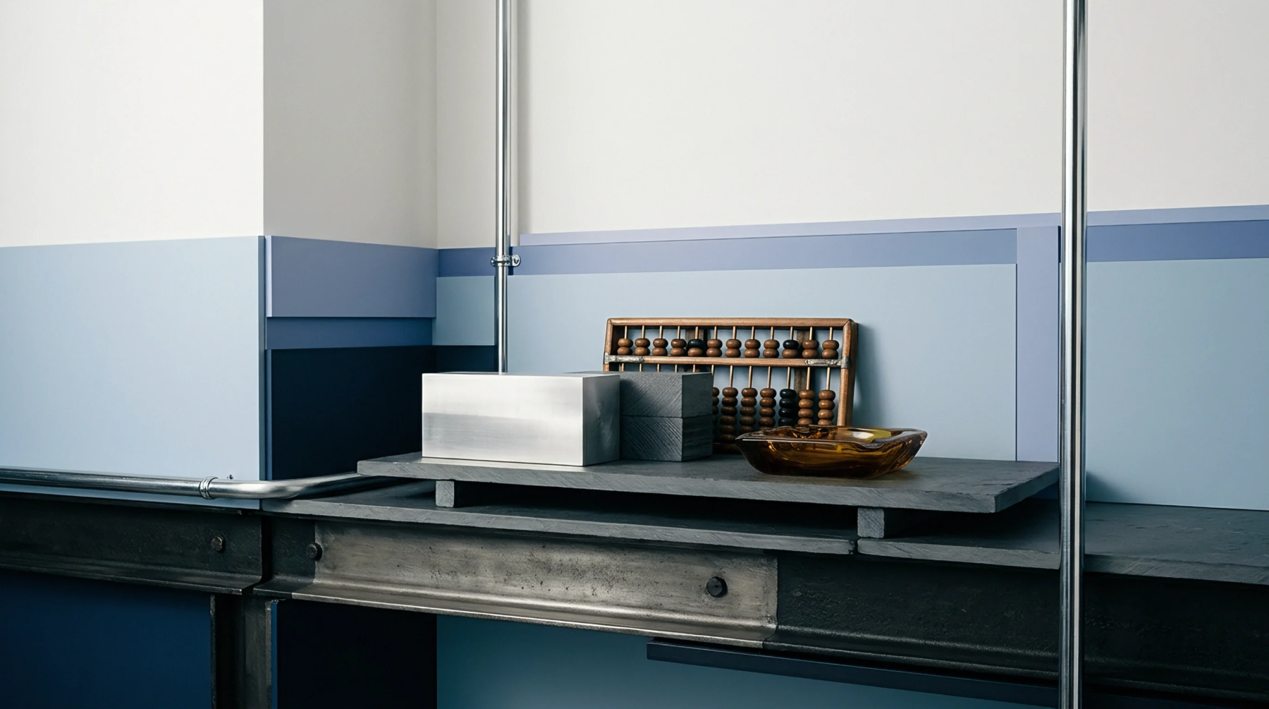 An abacus steel blocks and a glass dish sit on a grey stone slab atop a metal worktable framed by painted walls.