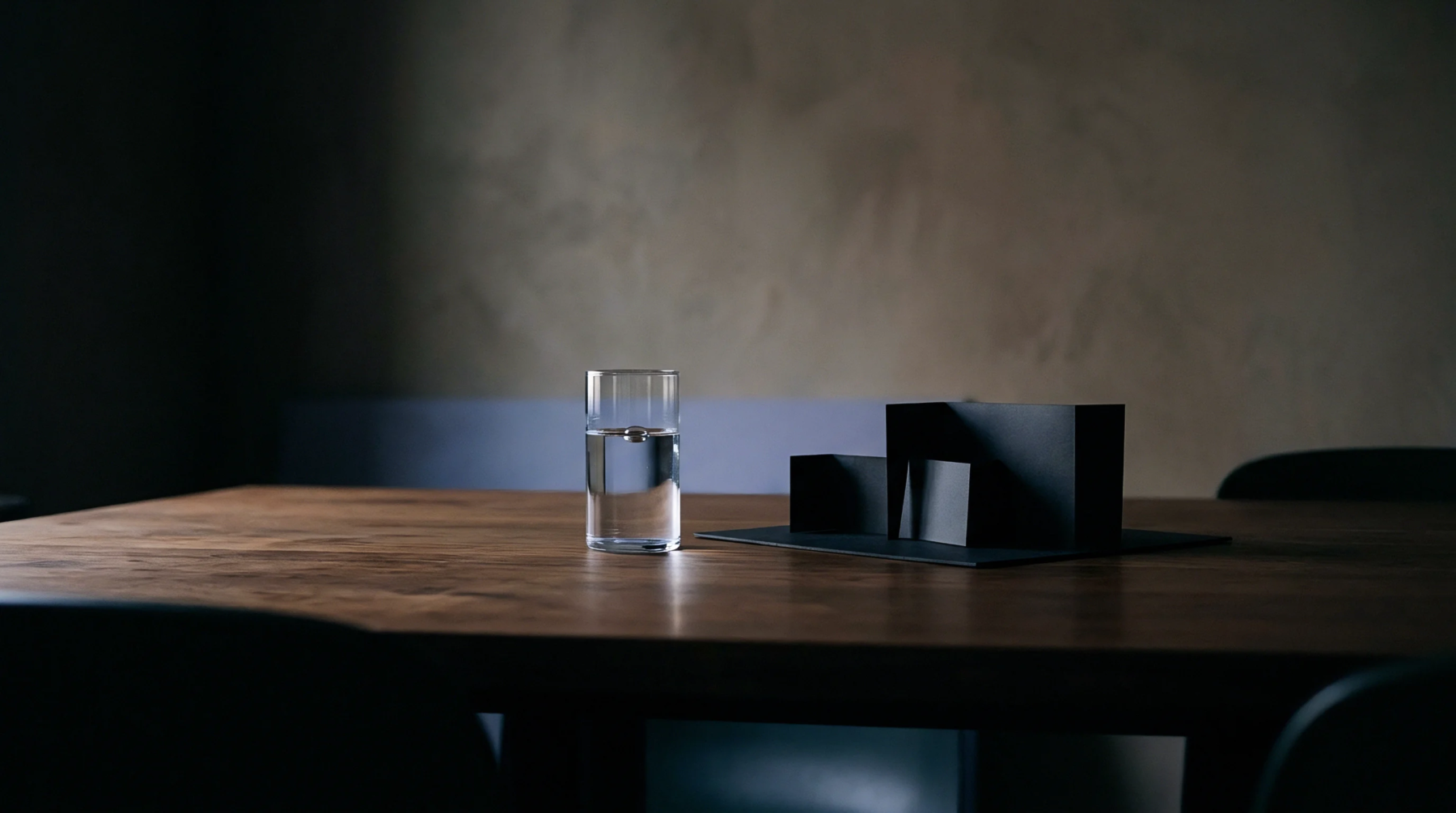 A clear glass of water stands beside several black geometric models arranged on a dark wooden table in a dimly lit industrial design setting.
