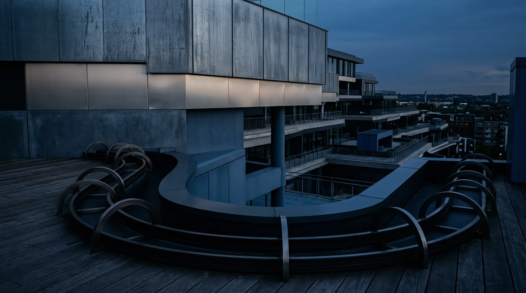 Modern building architecture with dark metallic panels and tiered balconies adjoins a low, serpentine installation on a wooden rooftop deck.