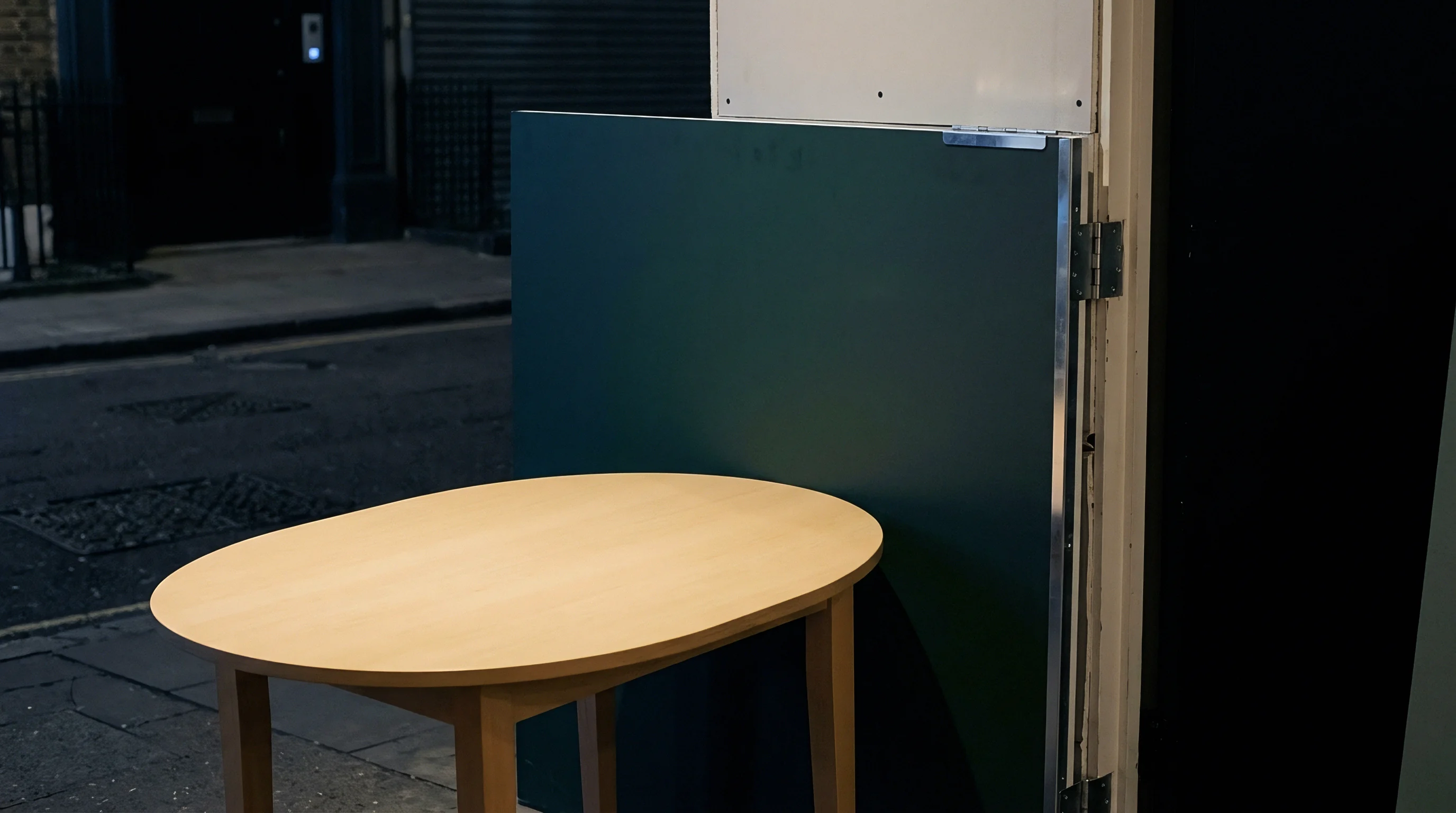 An oval table sits before a green modular partitioning system positioned on a paved exterior surface during twilight hours.