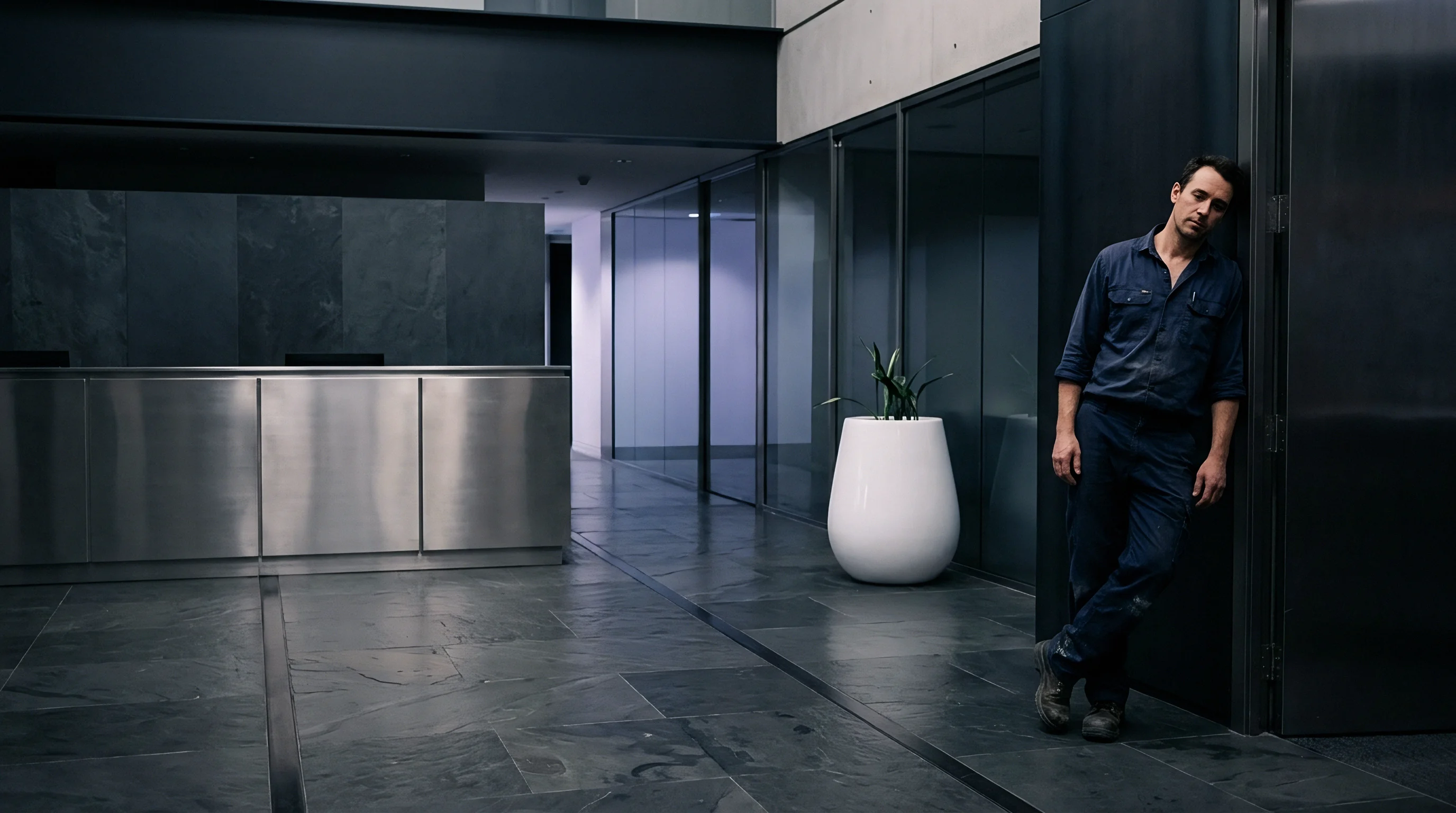 A worker in blue utility coveralls leans against a dark interior wall near a stainless steel service desk and white floor planter.