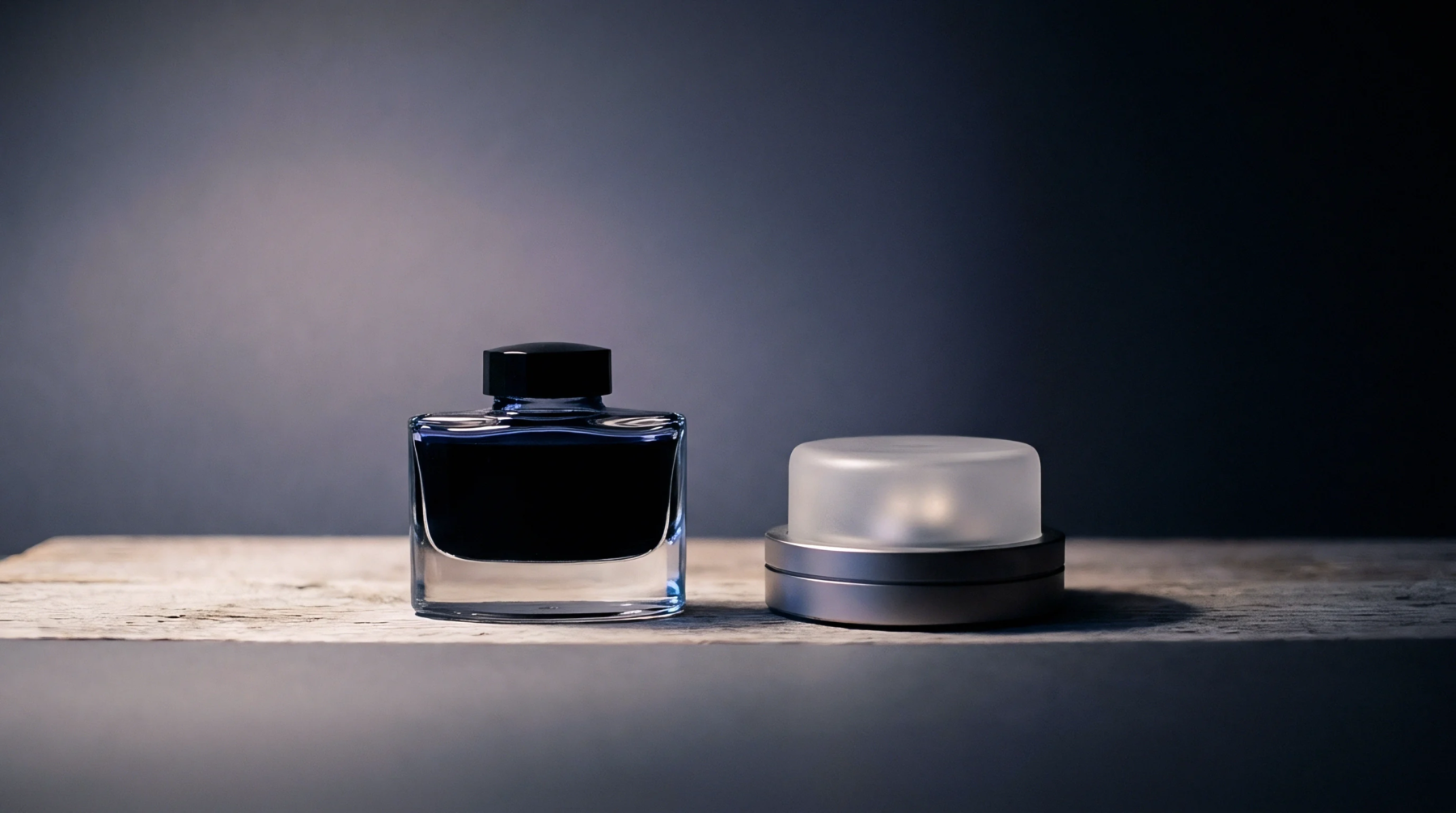 A square glass ink vessel sits beside a circular metallic housing component on a rustic wooden workbench against a dark industrial backdrop.